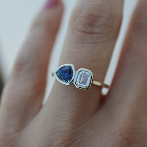 Könnte beinhalten: Ein goldener Ring mit einem blauen dreieckigen Saphir und einem klaren rechteckigen Diamanten.