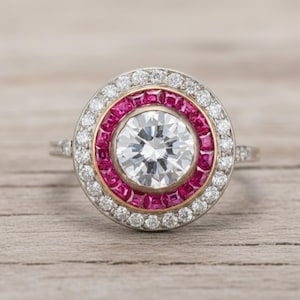 Könnte beinhalten: Ein runder, weißer Diamant-Verlobungsring mit einem Halo aus kleinen Diamanten und einem Ring aus roten Edelsteinen. Der Ring hat ein goldenes Band.