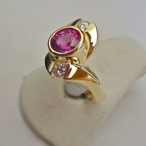 May include: A gold ring featuring a large, oval-cut pink gemstone as the centerpiece. The ring has a unique design with two smaller diamonds and gold accents. The ring is set on a neutral-colored display.
