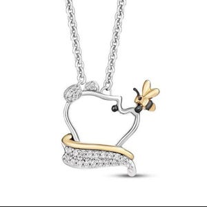 Może przedstawiać: Srebrny naszyjnik z wisiorkiem w kształcie słoika miodu. Słoik miodu ma złote akcenty i jest inkrustowany małymi, przezroczystymi kamieniami. Mała, złoto-czarna pszczoła siedzi z boku słoika miodu.