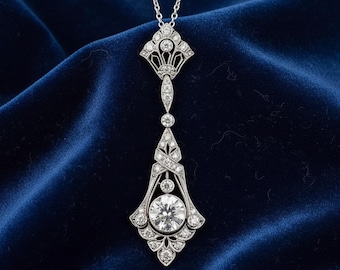 May include: A silver pendant necklace with a detailed Art Deco design. The pendant features a large, round, clear gemstone at the center, surrounded by smaller stones and intricate metalwork. The necklace hangs on a delicate silver chain against a dark blue background.