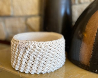 May include: A white crocheted bowl with a small white label that says "The" on it. The bowl is sitting on a light brown surface.