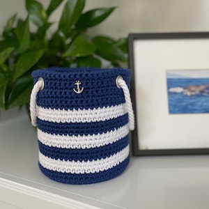 May include: A blue and white striped crocheted storage basket with a silver anchor charm. The basket has white rope handles.