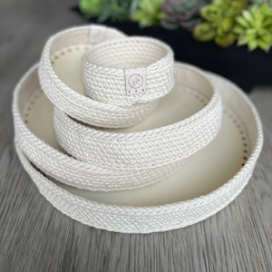 May include: Three white, round, woven rope baskets of varying sizes, stacked on top of each other. The smallest basket has a brown label with the word "smile" on it.