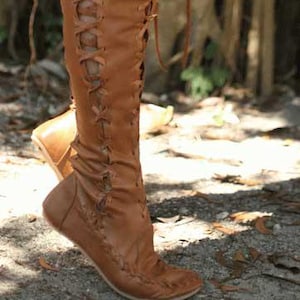 Puede incluir: Botas altas de cuero marrón con diseño de cordones. Las botas tienen suela plana y están hechas de cuero suave y flexible. Los cordones suben por la parte delantera de las botas y la parte superior de las botas es ligeramente acampanada.