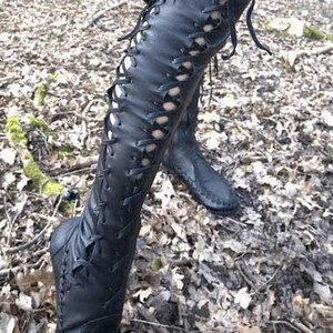 Peut inclure: Une paire de bottes noires montant jusqu'au genou, dot&eacute;es d'un la&ccedil;age frontal sur toute la longueur avec de fins lacets noirs. Les bottes pr&eacute;sentent des coutures visibles sur les bords et une structure souple remontant jusqu'au genou.