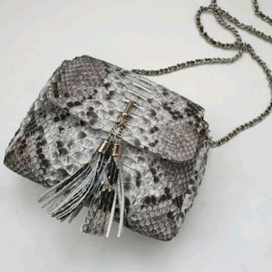 Op de afbeelding: Een kleine, rechthoekige handtas met een slangenpatroon in grijstinten en wit. De tas heeft een zilveren kettingriem en een kwastdetail. De klepsluiting is beveiligd met een zilveren sluiting.
