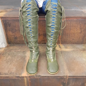 Peut inclure: Bottes vert olive montant jusqu'aux genoux avec un la&ccedil;age frontal sur toute la longueur, des lacets assortis, des fermetures &eacute;clair lat&eacute;rales et des coutures d&eacute;coratives autour des orteils. Les bottes ont une semelle plate et couvrent les jambes.