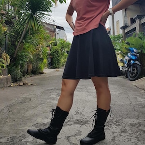 May include: A dusty rose short-sleeved t-shirt paired with a solid black knee-length skater skirt and black mid-calf lace-up boots. The skirt features a flared silhouette, while the boots have a rounded toe and black laces tied at the front.