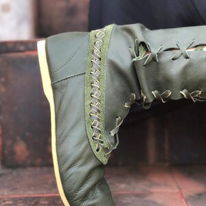 Peut inclure: Une botte en cuir vert olive dot&eacute;e d'un panneau lat&eacute;ral &agrave; la finition textur&eacute;e fa&ccedil;on daim et d'un motif d&eacute;coratif en point de croix. La botte pr&eacute;sente un la&ccedil;age avec des lacets fins de couleur claire et une semelle plate claire.