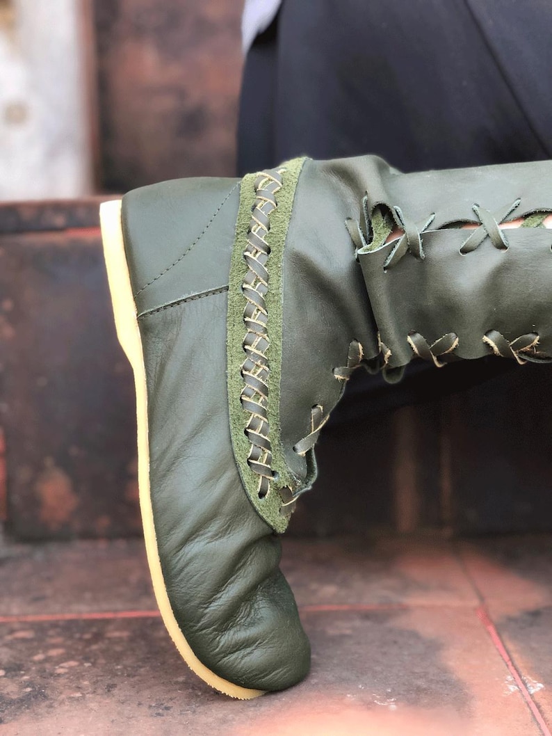 Peut inclure: Une botte en cuir vert olive dot&eacute;e d'un panneau lat&eacute;ral &agrave; la finition textur&eacute;e fa&ccedil;on daim et d'un motif d&eacute;coratif en point de croix. La botte pr&eacute;sente un la&ccedil;age avec des lacets fins de couleur claire et une semelle plate claire.