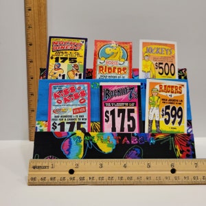 Può includere: Un porta-schede di tessuto nero con disegni di farfalle colorate. Il porta-schede ha slot per più biglietti della lotteria. I biglietti sono per giochi come "Nacho Dinero", "Kiss Kiss", "Rockin' 7's", "Horse Riders", "Jockeys" e "Racers".