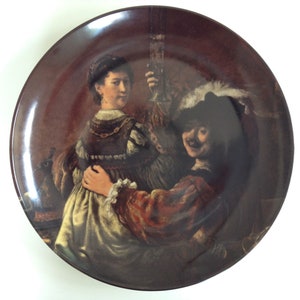 May include: A ceramic plate with a painting of two people, one holding a glass of wine. The painting is in a dark brown color scheme and the people are dressed in 17th century clothing.