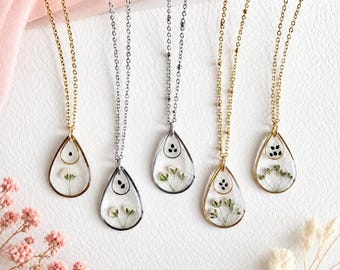 May include: Five teardrop-shaped pendant necklaces featuring pressed white flowers and small black seeds encased in clear resin. Three necklaces have gold-toned metal frames and chains, while two necklaces feature silver-toned metal frames and chains.