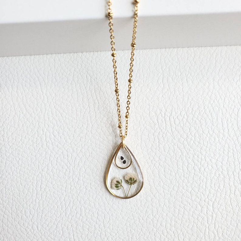 May include: A gold chain necklace with a teardrop-shaped pendant. The pendant is filled with clear resin and contains dried flowers and seeds.