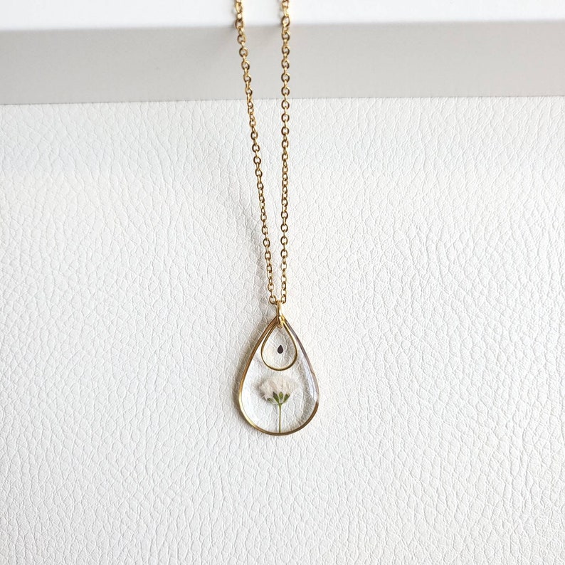 May include: A gold chain necklace with a teardrop-shaped pendant. The pendant is made of clear glass and contains a small white flower.