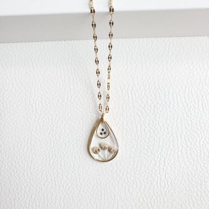May include: A gold chain necklace with a teardrop-shaped pendant. The pendant is made of clear resin and contains dried flowers and black seeds.