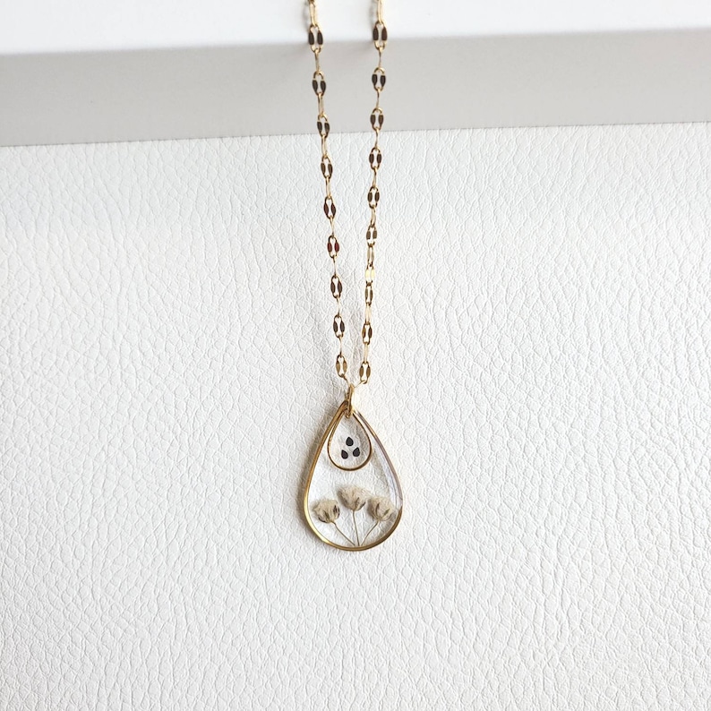 May include: A gold chain necklace with a teardrop-shaped pendant. The pendant is made of clear resin and contains dried flowers and black seeds.