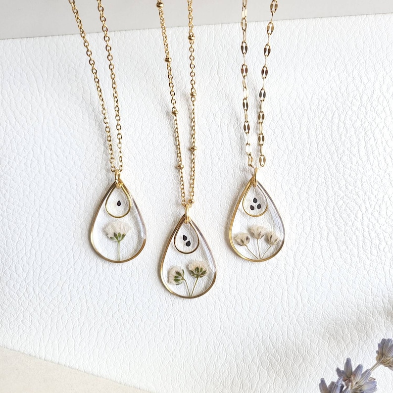 May include: Three gold necklaces with teardrop-shaped pendants featuring pressed flowers and black seeds inside clear resin.