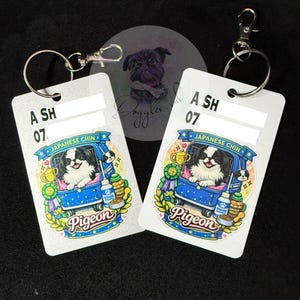 May include: Two rectangular white keychains with silver metal lobster clasps. Each features a colourful illustration of a Japanese Chin dog in a blue cart, with the text 'JAPANESE CHIN' and 'Pigeon', plus a trophy, shampoo, and treats.