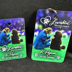 May include: Two rectangular tags featuring a photo of a person in a blue suit with a black poodle on a green surface. The text reads Lowashton Miniature Poodles, K Pegrum, and 07749 599908, accompanied by heart and paw print graphics.