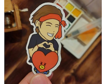 May include: A sticker featuring a cartoon character with brown hair and a red headband, forming a heart shape with their hands. The character is holding a red heart-shaped figure with a yellow face. A watercolor palette is in the background.