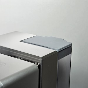 May include: A close-up of a silver coffee machine with a clear water tank and a grey lid. The lid has a textured surface.