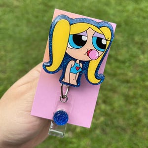 Könnte beinhalten: Ein Cartoon-Badge-Roller mit einer Figur mit gelbem Haar, blauen Augen und einer Kaugummiblase. Die Figur trägt ein blaues Oberteil mit einem Herz. Die Rolle hat eine rosa Rückseite und eine transparente Kunststoffrolle mit einem blauen Glitzerakzent.