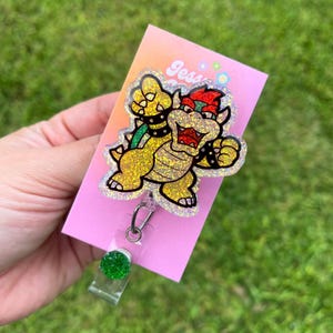 Può includere: Una carta rosa con il testo "Jess" su di essa. Un personaggio di Bowser scintillante dorato e verde è attaccato alla carta. Un porta badge glitterato verde è attaccato alla carta.