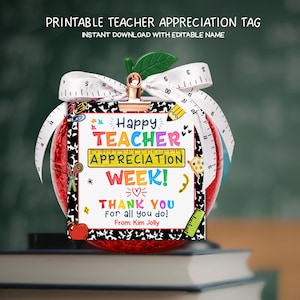May include: A red apple-shaped gift tag with a white ribbon and a gold clip. The tag reads "Happy TEACHER APPRECIATION WEEK! THANK YOU for all you do!" and is decorated with school-themed graphics. The tag is on top of a stack of books.
