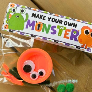 May include: A clear plastic bag containing craft supplies for making a monster. The bag includes an orange lid, googly eyes, orange and green pipe cleaners, and green pom-poms. The bag is labeled "MAKE YOUR OWN MONSTER" with colorful monster illustrations.