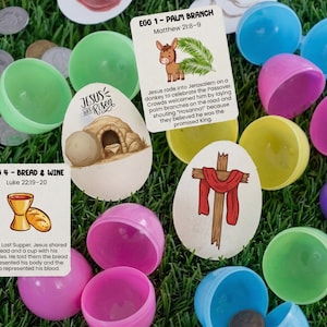 Op de afbeelding: Een assortiment kleurrijke plastic paaseieren, sommige open, verspreid op groen gras. Er zijn ook verschillende geïllustreerde kaarten met religieuze thema's, waaronder afbeeldingen van een ezel, een kruis en een graf.