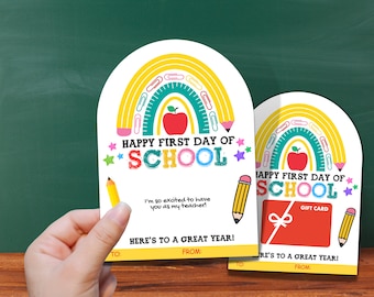 以下が含まれることがあります： 虹のデザイン、鉛筆、そして「Happy First Day of School」の文字が入った2つの白いギフトタグ。1つのタグにはギフトカードのグラフィックがあります。テキストは「I'm so excited to have you as my teacher!」と「Here's to a great year!」と書かれています。