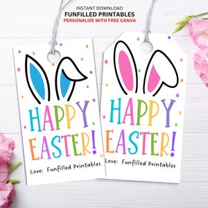 Puede incluir: Dos etiquetas de regalo imprimibles con orejas de conejo y el texto "¡Feliz Pascua!" en colores arcoíris. Las etiquetas están sobre un fondo de madera blanco.