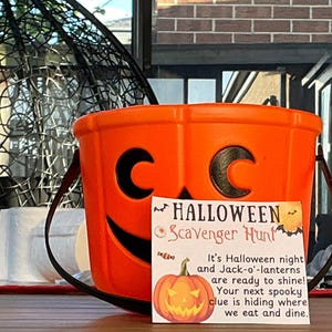 May include: An orange plastic Halloween bucket with a black handle and a jack-o'-lantern face. A sign in front reads "Halloween Scavenger Hunt" with a pumpkin illustration and text about Halloween night.