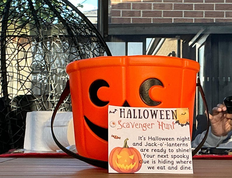 May include: An orange plastic Halloween bucket with a black handle and a jack-o'-lantern face. A sign in front reads "Halloween Scavenger Hunt" with a pumpkin illustration and text about Halloween night.