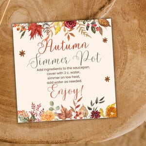 May include: A square card with the text "Autumn Simmer Pot" in elegant script, surrounded by watercolor illustrations of fall leaves, flowers, and spices. Instructions are included. The card is set on a wooden surface.