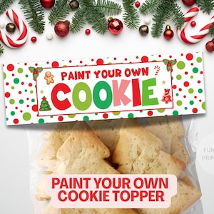 以下が含まれることがあります： ツリー型のクッキーが入った透明な袋で、「PAINT YOUR OWN COOKIE」と書かれたお祝いのバナーが付いています。バナーには赤、緑、白の水玉模様、ジンジャーブレッドマン、クリスマスツリーのイラストが描かれています。クリスマスオーナメントとモミの枝で飾られています。