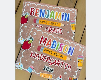 Può includere: Due cartelli rettangolari con sfondo marrone e bordi smerlati bianchi. I cartelli presentano testo colorato che recita Benjamin First day of 1st Grade e Madison First day of Kindergarten 2026, decorati con illustrazioni di mele, matite e materiale scolastico.