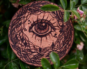May include: A round cork coaster featuring a black and white design of an eye surrounded by swirling lines. The coaster is resting on a bed of green leaves.