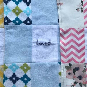 May include: A patchwork quilt with a variety of patterns in green, yellow, blue, pink, and white. The quilt has three white squares with the word "Loved" embroidered in black.