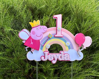 May include: A colorful cake topper featuring a cartoon pig character with a crown and fairy wings. The design includes a rainbow, the number "1", balloons, and the name "Jeyda". Perfect for a first birthday celebration.