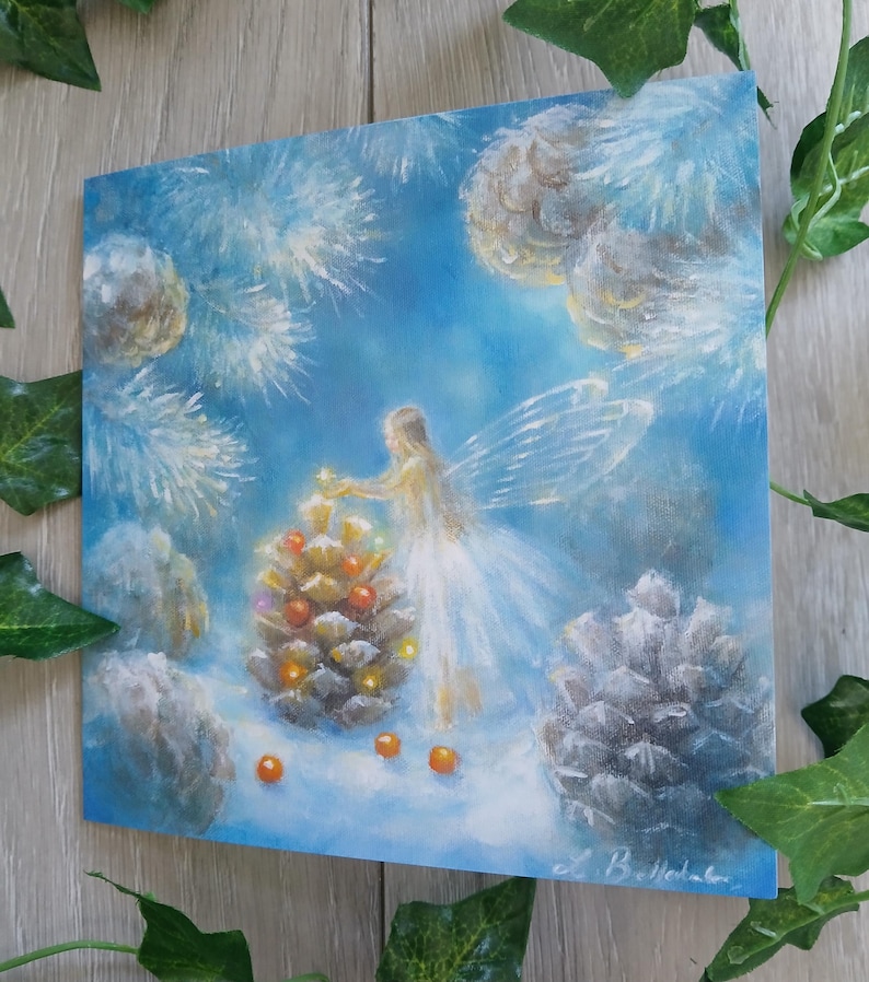 May include: An original painting of a fairy decorating a pinecone with orange ornaments. The fairy is wearing a white dress and has translucent wings. The background is a soft blue with white accents, and the painting is signed in the bottom right corner.