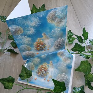 May include: Two greeting cards with a winter fairy theme. The cards feature a painting of a fairy decorating a pinecone with orange orbs against a blue sky with clouds and pine needles. The cards are blank inside.