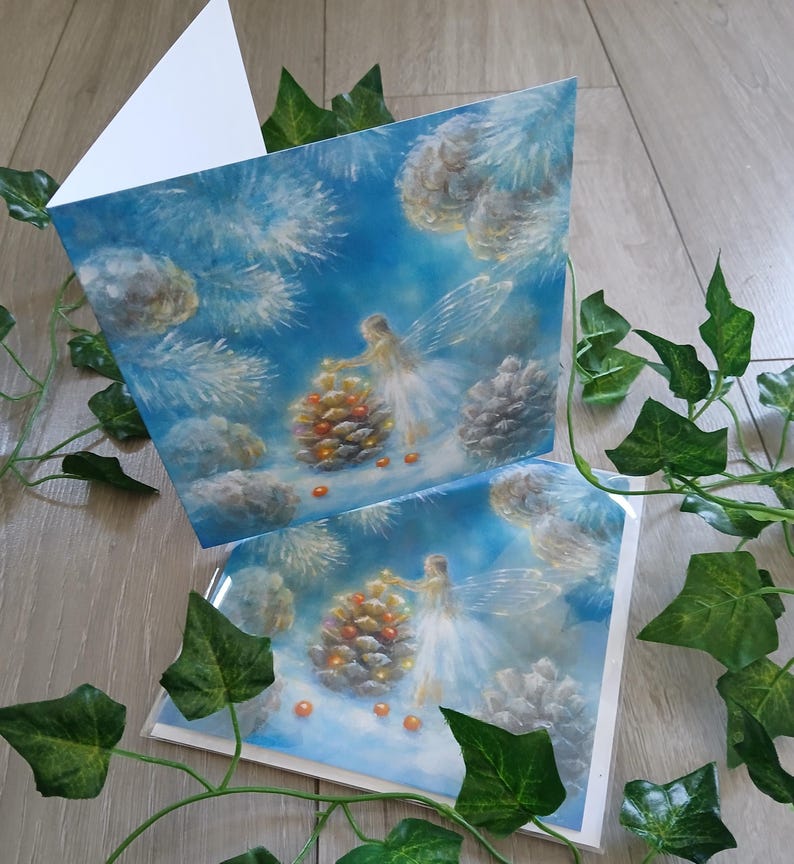 May include: Two greeting cards with a winter fairy theme. The cards feature a painting of a fairy decorating a pinecone with orange orbs against a blue sky with clouds and pine needles. The cards are blank inside.