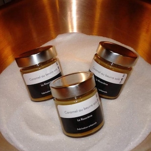 Puede incluir: Tres tarros de vidrio con tapas plateadas llenos de salsa de caramelo. Los tarros están etiquetados con "Caramel au beurre salé" y "La Ricocrème". Los tarros están en un tazón de cobre lleno de azúcar blanco.