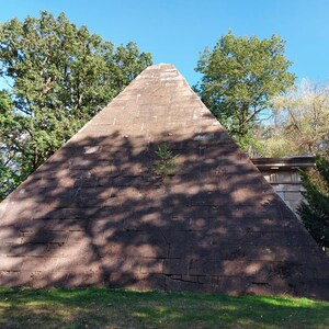 Könnte beinhalten: Eine große, verwitterte Steinpyramide, die aus rechteckigen Blöcken besteht. Die Oberfläche zeigt Altersspuren mit sichtbaren Mörtelfugen und einer kleinen grünen Pflanze, die aus der Mitte wächst. Die Pyramide steht vor Bäumen.