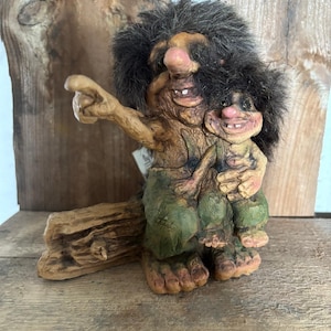 Könnte beinhalten: Eine skurrile Figur, die eine Trollfigur mit einem Kindertroll darstellt. Der erwachsene Troll hat eine große Nase, ein zahnreiches Grinsen und zeigt mit einem Arm. Der Kindertroll wird im Arm des Erwachsenen gehalten. Die Figuren sind in Erdtönen mit grüner Kleidung bemalt.