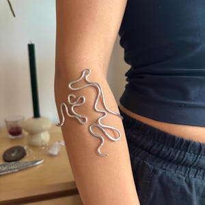 May include: A silver abstract arm cuff made of twisted wire. The cuff is worn on the upper arm, just below the elbow.