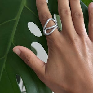 Könnte beinhalten: Ein silberfarbener Ring mit einem einzigartigen, offenen Design. Der Ring hat eine geschlungene Form, die eine abstrakte und moderne Ästhetik erzeugt. Der Ring wird auf einer Hand vor dem Hintergrund eines großen grünen Blattes gezeigt.